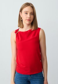 Top rosso senza maniche con dettaglio annodato sulla spalla sinistra, in tessuto morbido e dal taglio morbido, abbinato a jeans blu.