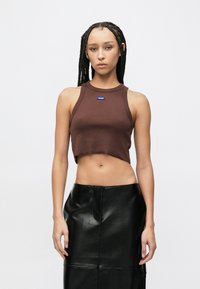 Καφέ κοντό τοπ με ριμπ ύφανση και στρογγυλή λαιμόκοψη με μπλε λογότυπο, συνδυασμένο με μαύρη φούστα από ψεύτικο δέρμα μήκους κάτω από το γόνατο, με σκίσιμο στο μπροστινό μέρος.