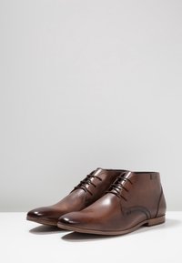 Stivali alla caviglia in pelle marrone con un design elegante, punta arrotondata, chiusura con lacci a sei occhielli e suola di un marrone chiaro a contrasto. Presenta cuciture sottili.