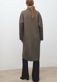 Lange bruine jas van zacht stof, met een rechte snit, detailnaden aan de achterkant en lange mouwen. Gecombineerd met zwart wijduitlopende broek.