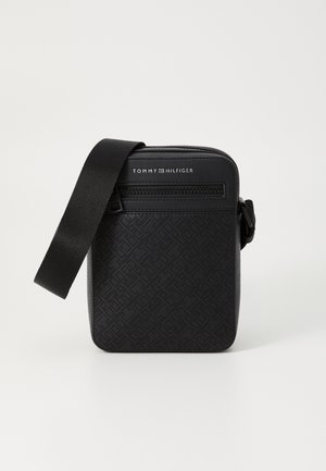 Czarna torba na ramię Tommy Hilfiger z wytłaczanym wzorem logo, przednią kieszenią na suwak i regulowanym paskiem na ramię na jednolitym tle.