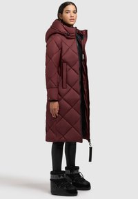 Burgunderfarbene gesteppte lange Jacke mit Kapuze, diagonalem Steppmuster, zwei Seitentaschen und Reißverschluss. Kombiniert mit schwarzen Leggings und Stiefeln.