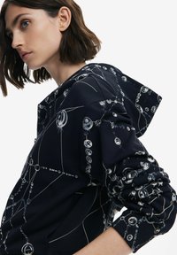 Sudadera marina con un estampado geométrico en azul claro y blanco. Los detalles incluyen una capucha con cordón y mangas fruncidas para darle textura.