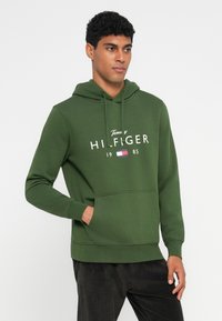 Hoodie vert en mélange de coton, avec une poche avant et une capuche à cordon. Texte logo blanc et année imprimés à l'avant.