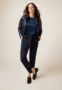 Donna in posa con giacca a vento blu scuro, top e pantaloni abbinati, un maglione annodato in vita e mocassini neri con motivo marrone.