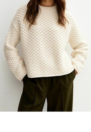 Femme portant un pull en tricot crème texturé associé à un pantalon ample vert olive foncé, les mains dans les poches.