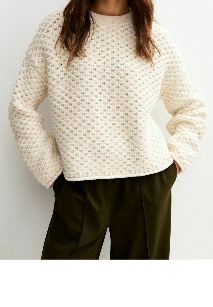 Femme portant un pull en tricot crème texturé associé à un pantalon ample vert olive foncé, les mains dans les poches.