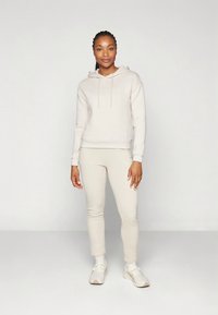 Felpa con cappuccio in pile beige con coulisse, abbinata a pantaloni tapered coordinati e scarpe da ginnastica bianche, caratterizzata da una texture liscia.