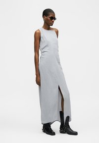 Femeie purtând o rochie lungă gri deschis fără mâneci, cu o deschizătură frontală, asortată cu cizme negre până la mijlocul gambei și ochelari de soare închiși la culoare, stând lateral.