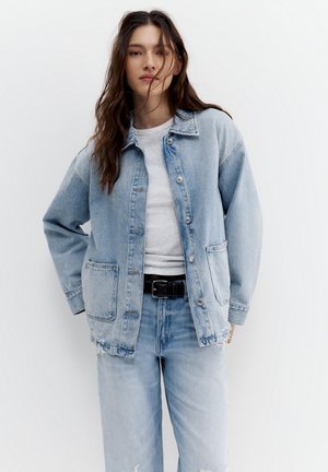 Jeune femme portant une veste en jean bleu clair et un jean avec une chemise blanche et une ceinture noire, posant avec une main dans la poche.