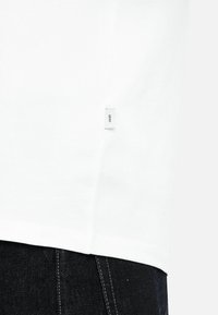 Weißes Kleidungsstück mit einem kleinen Stofflabel mit der Aufschrift "Orim" an der Seitennaht, getragen über dunklen Jeans mit sichtbaren Nähten.