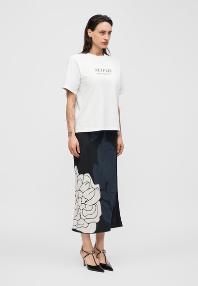 Γυναίκα που στέκεται φορώντας λευκό t-shirt με γραφικό της Netflix, μαύρη και λευκή φλοράλ μίντι φούστα και μαύρα μυτερά τακούνια με λουράκια στον αστράγαλο.