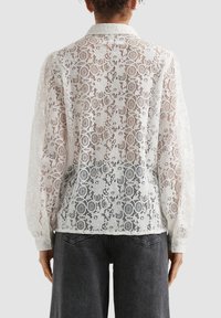 Woman wearing a white long-sleeve lace blouse with floral patterns, paired with dark gray denim jeans, viewed from the back.