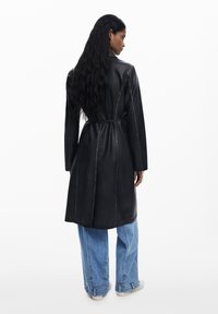 Cappotto lungo in pelle nera con vita cintata, caratterizzato da cuciture visibili e un taglio dritto, abbinato a jeans in denim blu larghi.