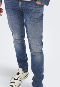 Jeans in denim di blu medio con una finitura sbiadita e leggere abrasioni; abbinati a sneakers bianche e nere con dettagli verdi.