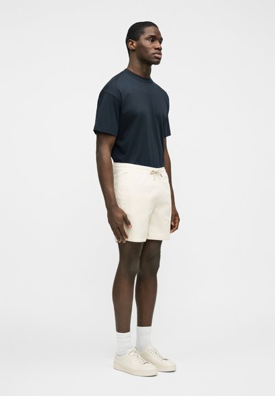 Homme debout de profil portant un t-shirt noir, un short beige, des chaussettes blanches et des baskets beiges sur un fond blanc uni.