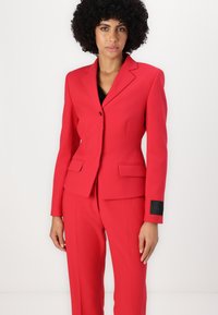 Blazer ajusté rouge avec revers crantés, fermeture à un bouton, deux poches avant et accent de label noir sur le poignet. Texture de tissu lisse.