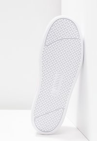 Semelle de basket blanche avec motif d’adhérence texturé et logo centré, positionnée à la verticale contre un coin blanc du mur et du sol.