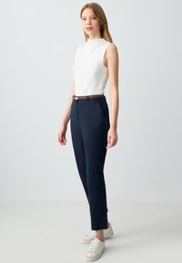 Jonge vrouw in een mouwloos wit shirt, marineblauwe broek met een bruine riem, en witte sneakers staand tegen een eenvoudige lichte achtergrond.