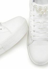 Witte sneakers met een gladde leren buitenkant. Kenmerken zijn platte veters en een decoratieve rij parels op de bovenkant. Gestructureerde rubberen zool.