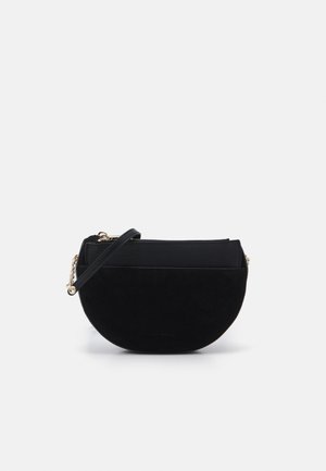 Sort crossbody-taske i halvmåneform med læderrem og guldkædedetaljer, sat mod en ensfarvet lys baggrund.