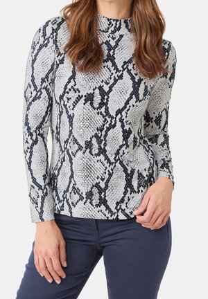 Femme portant un haut à manches longues gris et noir à motif peau de serpent avec un pantalon bleu foncé, debout avec une main sur la hanche.