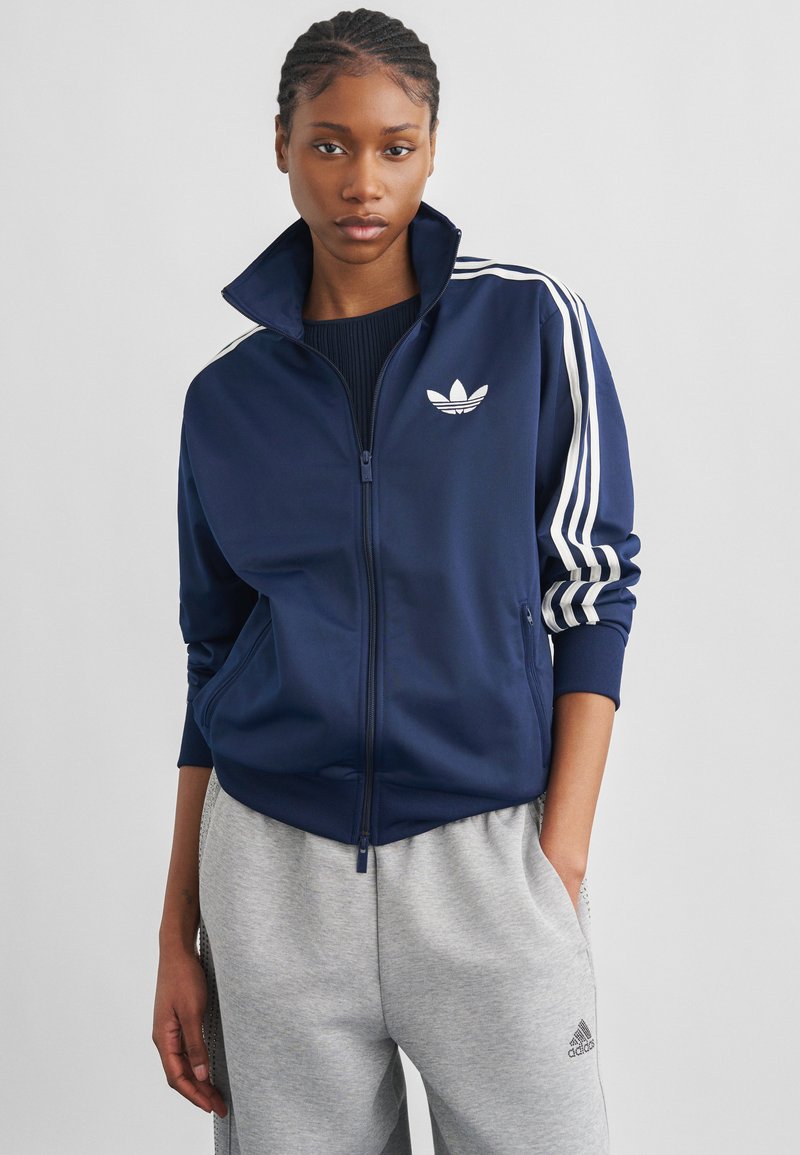 Junge Frau, die eine marineblaue Adidas-Jacke mit Reißverschluss und weißen Streifen sowie graue Adidas-Sweatpants trägt, steht mit einer Hand in der Tasche.