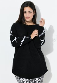 Frau mit dunklen Haaren, die einen schwarzen Strickpullover mit weißen Schleifenakzenten an den Ärmeln und schwarzen mit weißen Mustern dekorierten Hosen trägt.