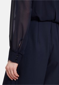 Marinblus med genomskinliga långa ärmar, knapddetaljer vid manschetterna och en slät, texturerad kropp. Huvudfokus på färg- och tygkombination.