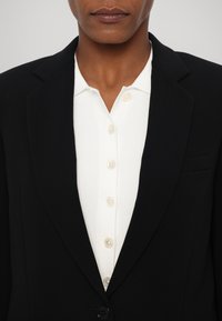 Svart blazer med spiss krage over en hvit ribbet skjorte med knapper. Har klare knapper og en brystlomme på blazeren.