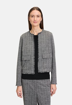 Vrouw in een grijs jasje met visgraatpatroon en bijpassende broek over een zwarte top, staande tegen een effen lichte achtergrond.