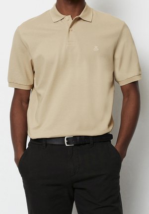 Mann trägt beige kurzärmliges Polohemd mit kleinem weißem Logo, schwarzen Gürtel und schwarze Hose, Hände in den Taschen vor einfarbigem Hintergrund.