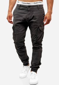 Cargo byxor i mörkgrått tyg med elastiska muddar, flera sidofickor och en midjeband med Calvin Klein-logotyp. Bärs med vita sneakers.
