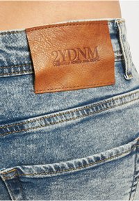 Denimjeans med en brun lærflekk med preget tekst på linningen. Lyseblått stoff med falming og kontrasterende gul søm.