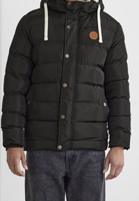 Schwarze gesteppte Jacke mit vorderen Knöpfen, zwei Seitentaschen, weißen Kordeln an der Kapuze, getragen mit dunkelgrauen Jeans, gezeigt von den Schultern bis zur Taille.