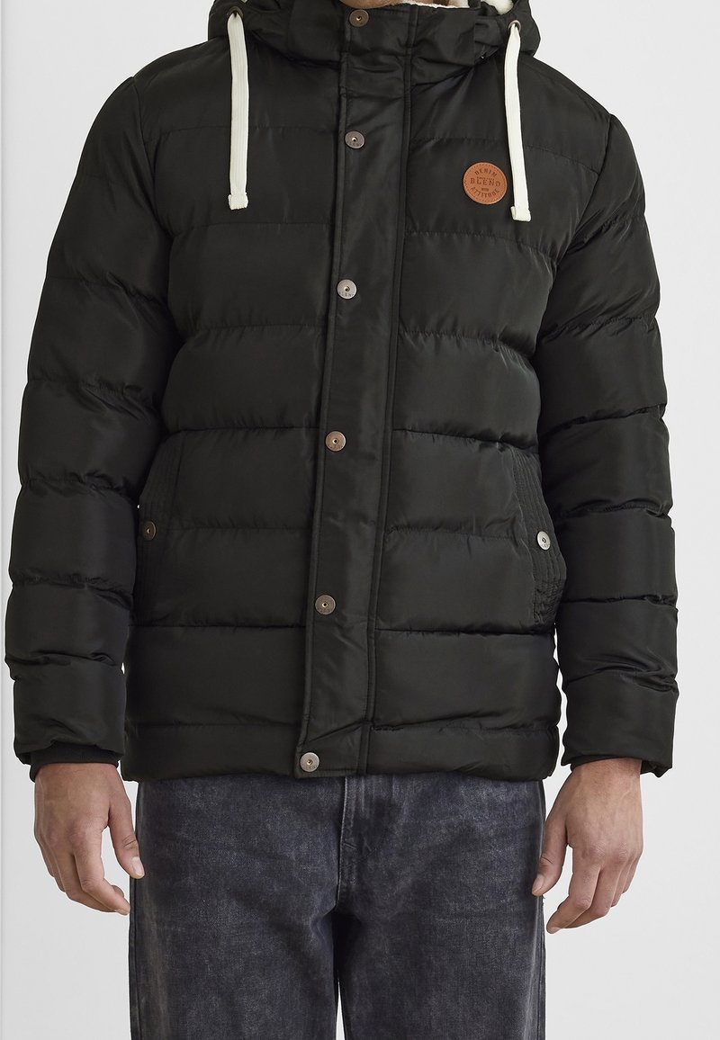 Schwarze gesteppte Jacke mit vorderen Knöpfen, zwei Seitentaschen, weißen Kordeln an der Kapuze, getragen mit dunkelgrauen Jeans, gezeigt von den Schultern bis zur Taille.