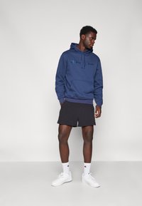 Felpa con cappuccio blu navy con tasca frontale e lacci, abbinata a pantaloni corti neri e sneakers bianche. Il logo Champion è ben visibile.
