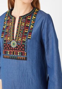 Mujer con una blusa de manga larga azul con un patrón geométrico colorido bordado alrededor del escote con abertura.