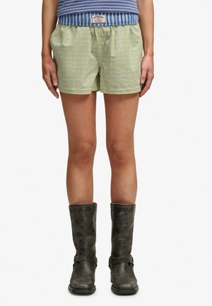 Osoba nosząca żółto-niebieskie szorty w kratę, niebiesko-biały pasiasty top oraz ciemne kozaki do połowy łydki z detalami sprzączek.