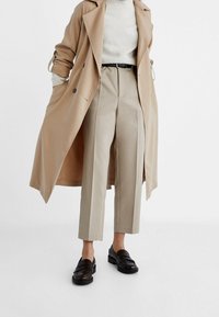 Beige trenchcoat over een witte trui, gecombineerd met hooggewaist beige broek en donkere instappers. Eenvoudig ontwerp, gladde textuur, neutraal palet.
