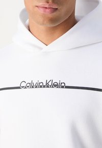 Hanorac alb cu o linie orizontală neagră și logo-ul "Calvin Klein". Material moale cu glugă cu șnur și detalii vizibile de coasere.