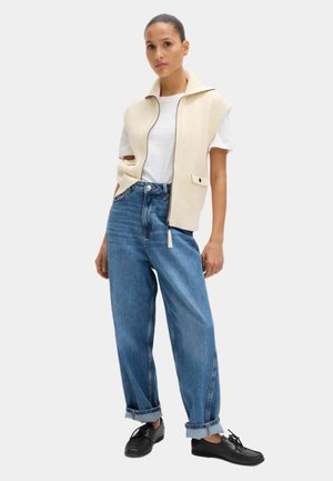 Gilet senza maniche beige a coste con colletto, indossato sopra una t-shirt bianca e jeans blu a taglio largo, abbinato a scarpe nere con lacci.