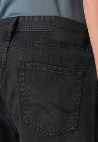 Jeans negre din denim cu o suprafață texturată, având un buzunar pe spate și o etichetă de marcă rectangulară. Accentele de cusătură subtile sunt vizibile.