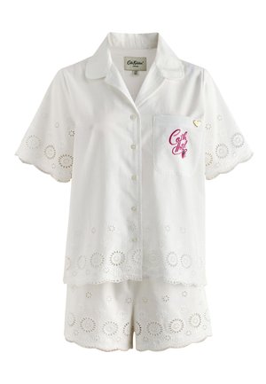 Vrouw in wit ooglet kort mouwen shirt en bijpassende short staat blootsvoets op een balkon met een schilderachtige heuvel en gebouwen op de achtergrond bij zonsondergang.