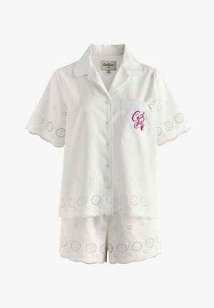Vrouw in wit ooglet kort mouwen shirt en bijpassende short staat blootsvoets op een balkon met een schilderachtige heuvel en gebouwen op de achtergrond bij zonsondergang.