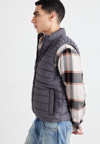 Chaleco gris acolchado con cuello alto, cremallera frontal y bolsillos laterales, superpuesto a una camisa de manga larga de cuadros en tonos beige y negro.