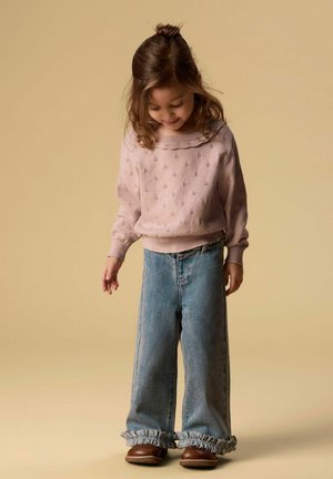 Noor tüdruk vaatab allapoole, kandes roosat mustrilist kampsunit, laia jalaga siniseid pükse rebenenud äärega ja pruune kingi beežil taustal.