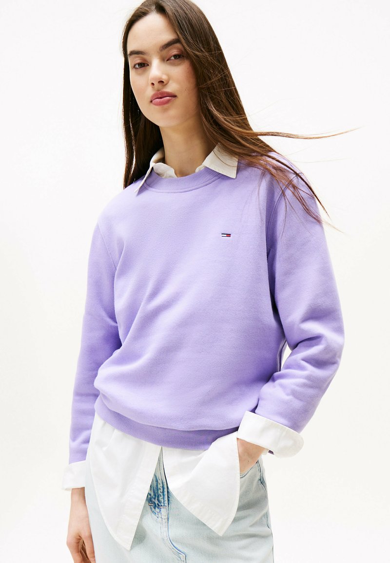 Sudadera lavanda con cuello y puños acanalados, usada sobre una camisa blanca con cuello. El suéter presenta un pequeño logo en el pecho.