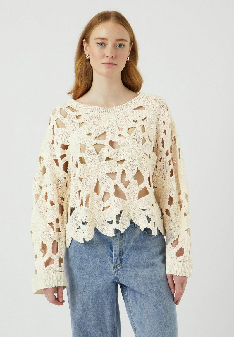 Maglione all'uncinetto color crema con motivo floreale, dal design a maglia aperta, maniche ampie e colletto a coste, abbinato a jeans di denim a vita alta.