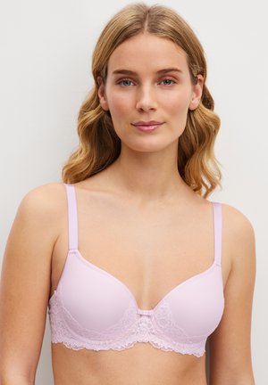 Jeune femme aux cheveux blonds portant un soutien-gorge rose clair à bordure en dentelle sur un fond clair uni.